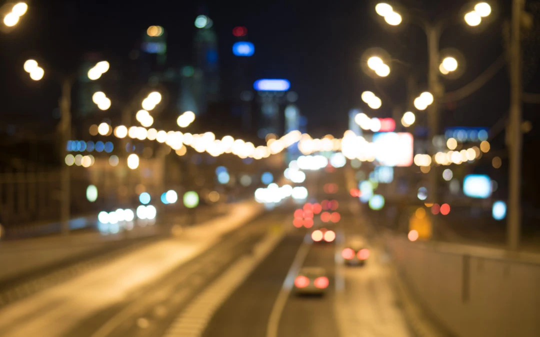 Blurry nighttime image of a city street illuminated by outdoor lighting, showcasing cars on the road and city skyline in the background, relevant to outdoor lighting protection tips from Kazar's Electric.