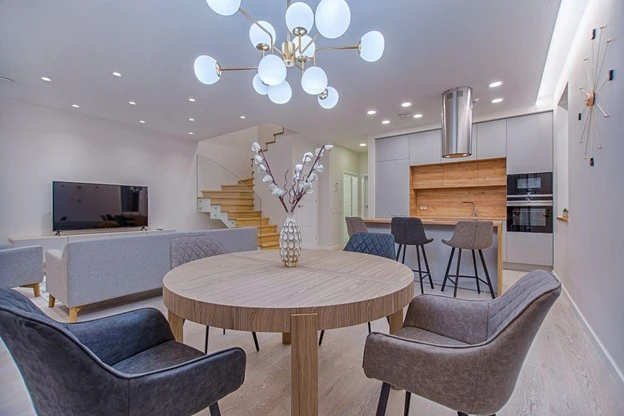 Modern living room with stylish lighting fixture, round wooden dining table, and comfortable chairs, showcasing an inviting ambiance and contemporary design elements.